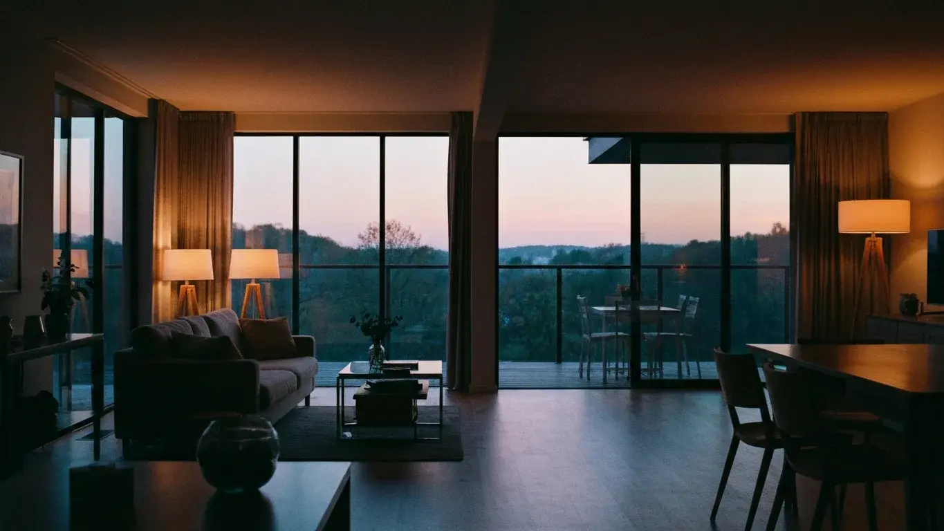 editorial interior vignette of a open-plan living room with floor-to-ceiling windows framing a nature view, lit by table lamps at dusk